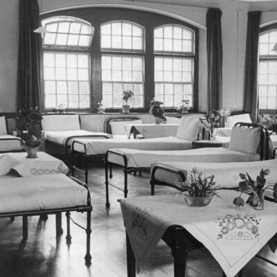 Dormitory beds at Glenside Hospital, with flowers decorating the tables and large windows in the ward