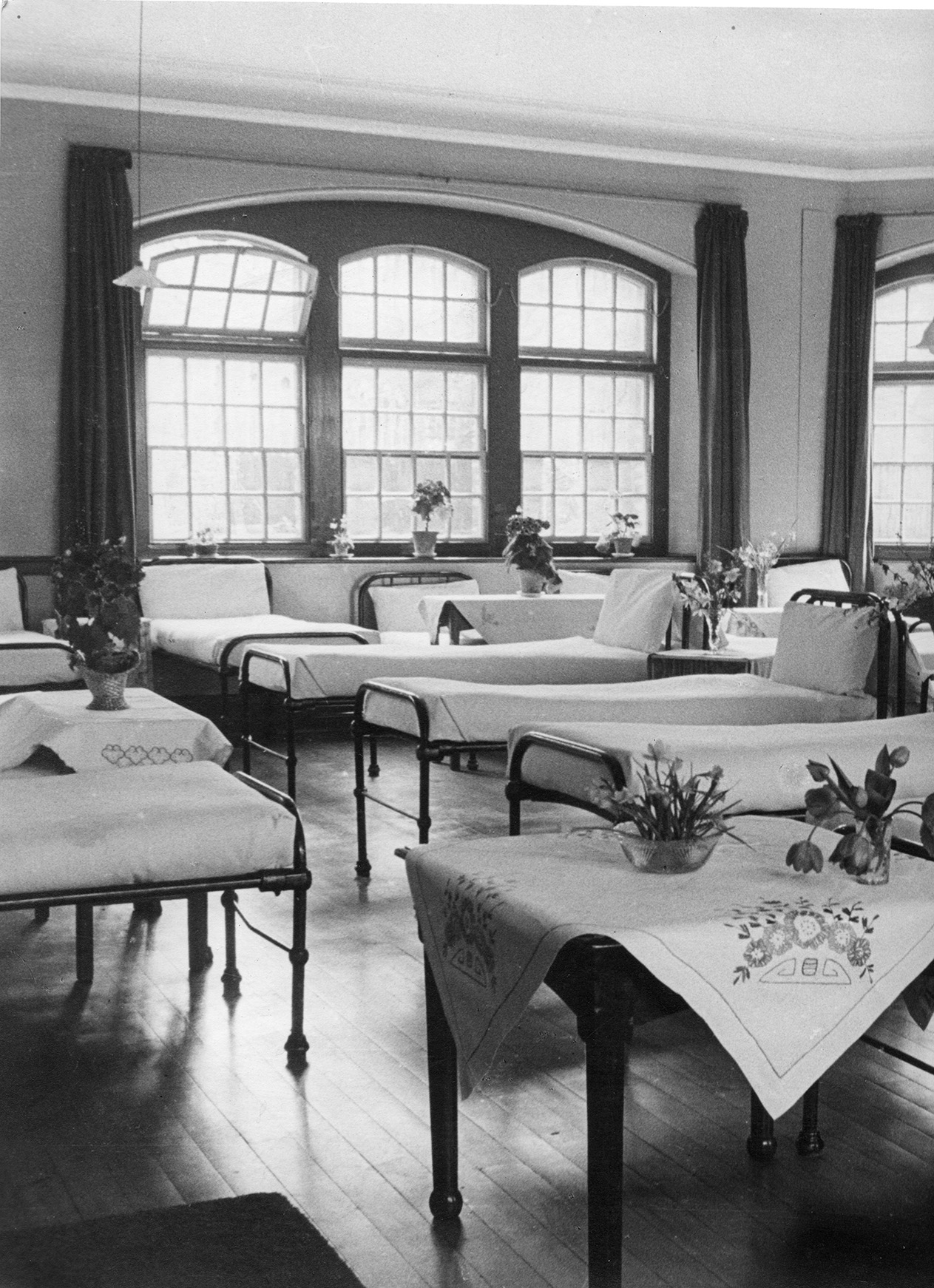 Dormitory beds at Glenside Hospital, with flowers decorating the tables and large windows in the ward